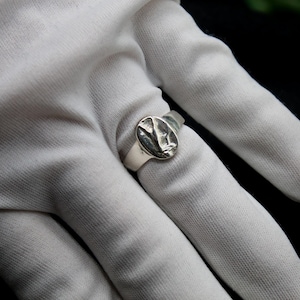 Pu&ograve; includere: Un anello a sigillo d'argento con un design ovale con un motivo testurizzato. L'anello &egrave; indossato su un dito con un guanto bianco.