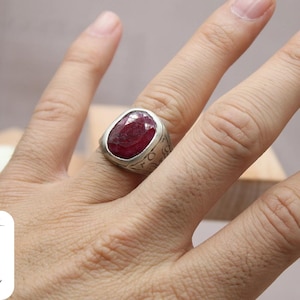 May include: A silver ring with a large, oval-shaped, red gemstone set in the center. The ring has intricate details around the gemstone.