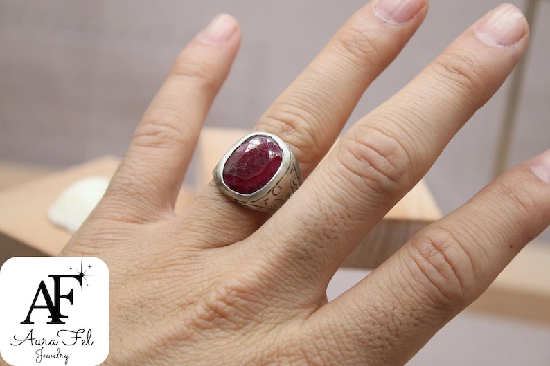 May include: A silver ring with a large, oval-shaped, red gemstone set in the center. The ring has intricate details around the gemstone.