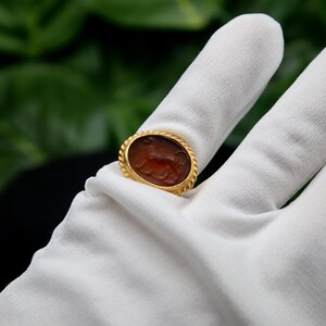 May include: A gold ring with a brown oval cameo featuring a carved design. The ring is on a white surface.