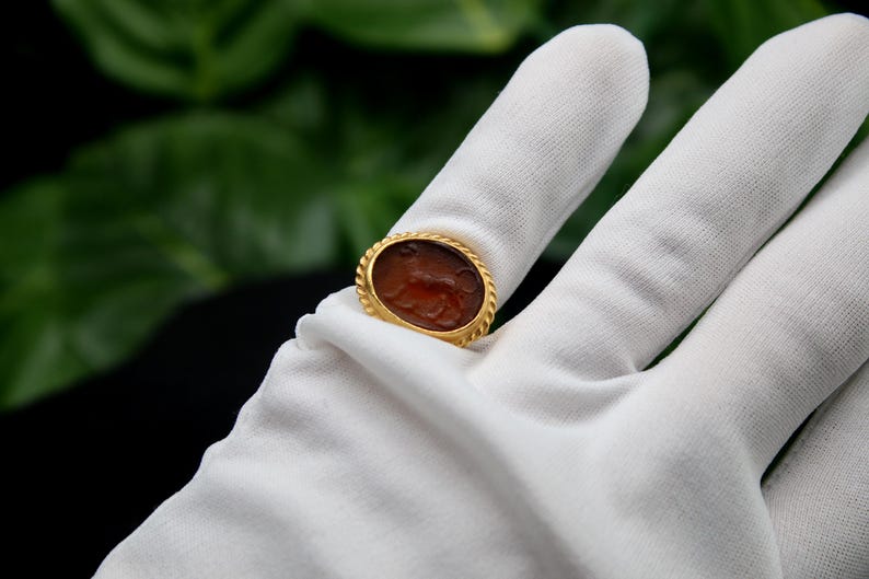 May include: A gold ring with a brown oval cameo featuring a carved design. The ring is on a white surface.