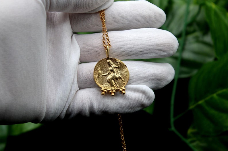 May include: A gold necklace with a round pendant featuring a detailed embossed image of a woman riding a horse. The pendant has a series of small gold beads hanging from the bottom.