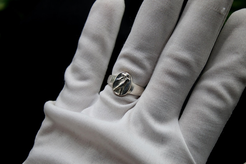 Pu&ograve; includere: Un anello d'argento con un design ovale che presenta un uccello stilizzato. L'anello &egrave; indossato su una mano guantata di bianco.