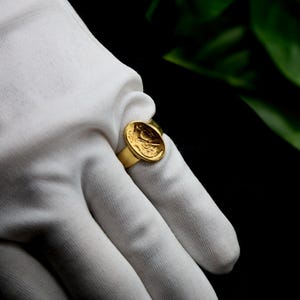 Pode incluir: Um anel de ouro com um design redondo em relevo que apresenta uma imagem detalhada de um p&aacute;ssaro. O anel &eacute; usado em uma m&atilde;o enluvada.