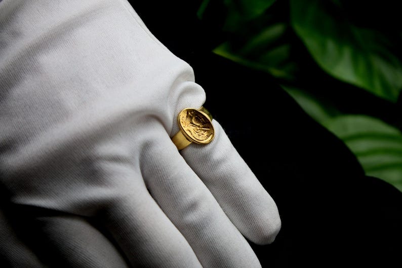 Pode incluir: Um anel de ouro com um design redondo em relevo que apresenta uma imagem detalhada de um p&aacute;ssaro. O anel &eacute; usado em uma m&atilde;o enluvada.