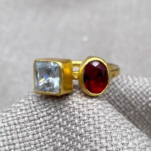 May include: A gold-toned ring featuring two distinct gemstones set side-by-side. One stone is a clear, square-cut gem, while the other is a deep red, oval-cut gem. The band has a textured, metallic finish, creating a bold and contrasting appearance.