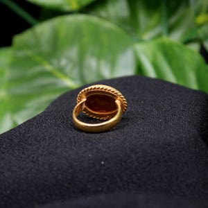 May include: A gold ring with a large oval-shaped stone set in a bezel setting. The ring is on a black fabric background.
