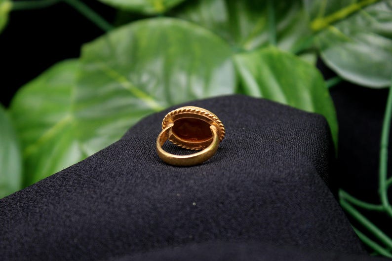 May include: A gold ring with a large oval-shaped stone set in a bezel setting. The ring is on a black fabric background.
