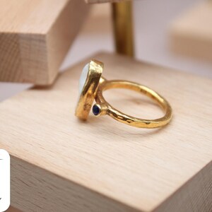 May include: A gold ring with a white oval gemstone and a small blue gemstone accent. The ring has a hammered texture.