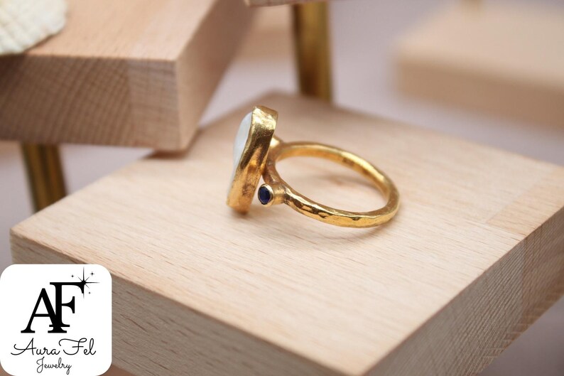 May include: A gold ring with a white oval gemstone and a small blue gemstone accent. The ring has a hammered texture.
