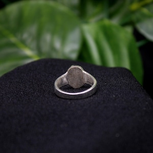 Pu&ograve; includere: Un anello d'argento con una parte superiore liscia di forma ovale. L'anello &egrave; su uno sfondo di tessuto nero.