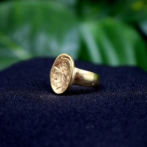 Pode incluir: Um anel de ouro com um design oval que apresenta uma imagem em relevo de uma cabe&ccedil;a humana. O anel est&aacute; sobre uma superf&iacute;cie preta.