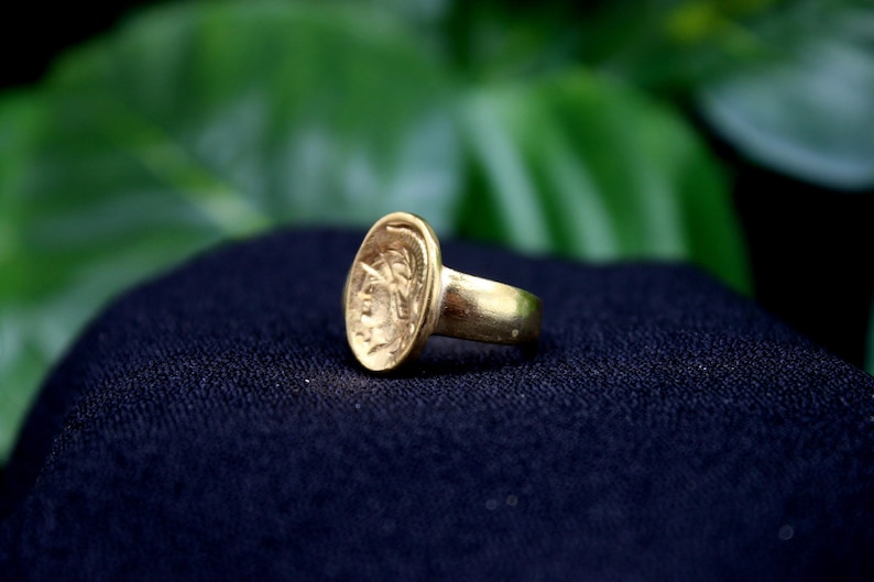 Pode incluir: Um anel de ouro com um design oval que apresenta uma imagem em relevo de uma cabe&ccedil;a humana. O anel est&aacute; sobre uma superf&iacute;cie preta.