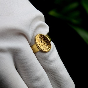 Pode incluir: Um anel de ouro com um design redondo em relevo que apresenta uma imagem detalhada do rosto de uma pessoa. O anel &eacute; usado em uma m&atilde;o enluvada de branco.