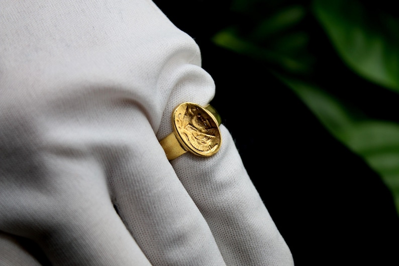 Pode incluir: Um anel de ouro com um design redondo em relevo que apresenta uma imagem detalhada do rosto de uma pessoa. O anel &eacute; usado em uma m&atilde;o enluvada de branco.