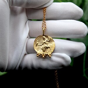 May include: A gold necklace with a round pendant featuring a detailed image of a woman holding a staff. The pendant has five small gold beads hanging from the bottom.