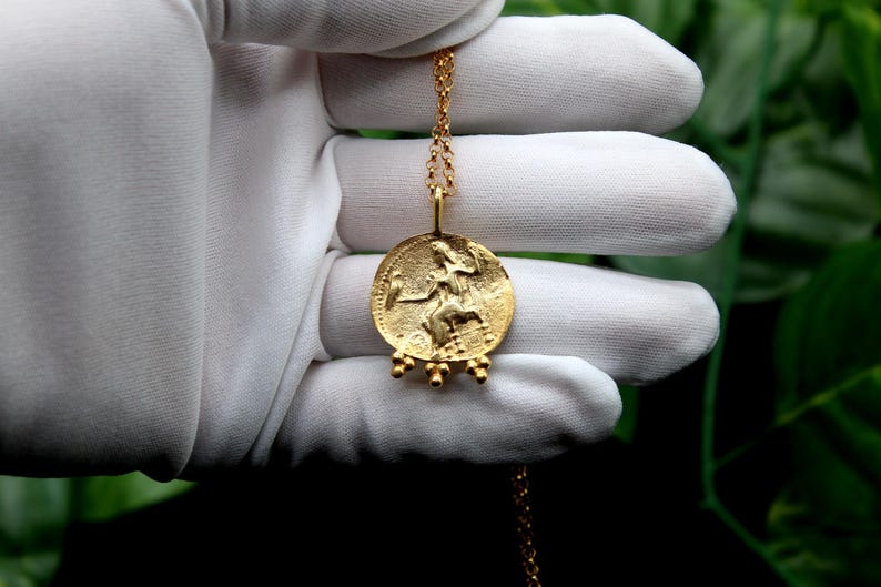 May include: A gold necklace with a round pendant featuring a detailed image of a woman holding a staff. The pendant has five small gold beads hanging from the bottom.