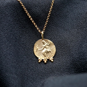 May include: A gold necklace with a round pendant featuring a detailed image of a woman holding a staff. The pendant has five small gold beads hanging from the bottom.