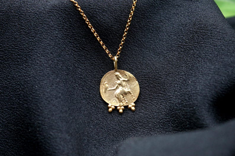 May include: A gold necklace with a round pendant featuring a detailed image of a woman holding a staff. The pendant has five small gold beads hanging from the bottom.