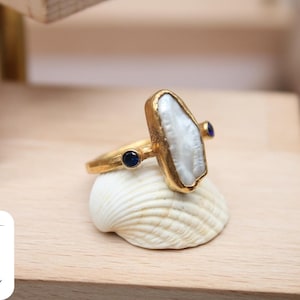 May include: A gold ring with a large white pearl-like stone and two small blue stones. The ring is displayed on a seashell.