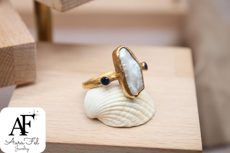 May include: A gold ring with a large white pearl-like stone and two small blue stones. The ring is displayed on a seashell.