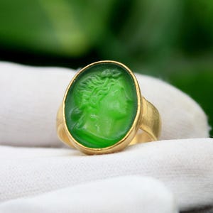 Può includere: Un anello in vetro verde con una fascia dorata. Il vetro presenta un design intagliato di un viso umano.