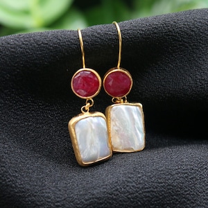 May include: Gold drop earrings featuring a red gemstone and a white, rectangular pearl. The earrings are suspended from a gold hook. The background is a black, textured fabric.