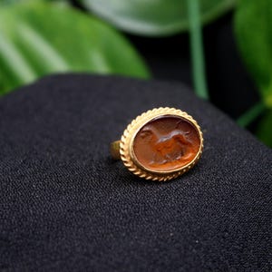 May include: A gold ring with an oval-shaped carnelian stone featuring a carved image of a lion. The ring is set on a black fabric background.