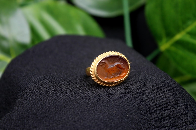 May include: A gold ring with an oval-shaped carnelian stone featuring a carved image of a lion. The ring is set on a black fabric background.