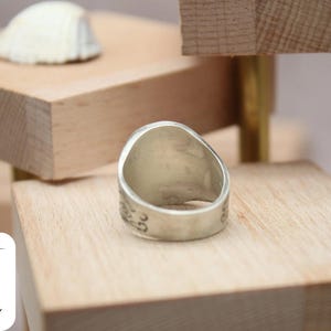 May include: A silver ring with a wide band and a smooth, rounded top. The ring is sitting on a wooden surface.
