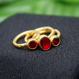 May include: Three gold rings with red gemstones. The rings are stacked on top of each other. The largest ring has a large oval-shaped red gemstone. The middle ring has a smaller round red gemstone. The smallest ring has a small round red gemstone.