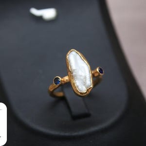 May include: A gold ring with a white pearl-like stone and two small blue stones. The ring is on a black display stand.