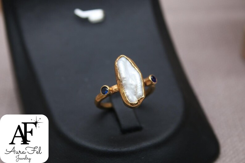 May include: A gold ring with a white pearl-like stone and two small blue stones. The ring is on a black display stand.