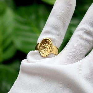 Pode incluir: Um anel de ouro com um design oval que apresenta um perfil detalhado de uma pessoa usando um capacete. O anel &eacute; exibido em uma superf&iacute;cie branca.