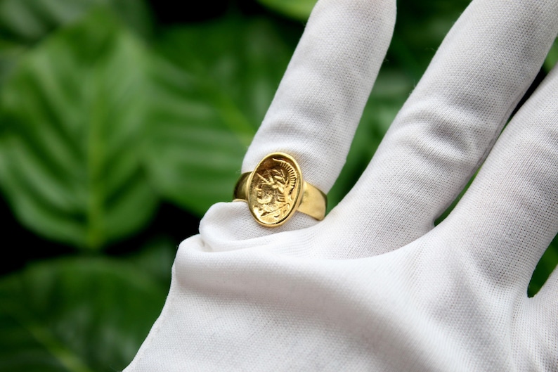 Pode incluir: Um anel de ouro com um design oval que apresenta um perfil detalhado de uma pessoa usando um capacete. O anel &eacute; exibido em uma superf&iacute;cie branca.