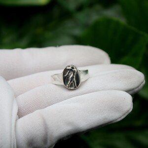 Pu&ograve; includere: Un anello d'argento con un design ovale che presenta una figura in rilievo di una persona. L'anello &egrave; mostrato su una superficie bianca.