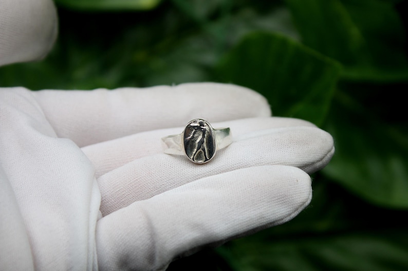Pu&ograve; includere: Un anello d'argento con un design ovale che presenta una figura in rilievo di una persona. L'anello &egrave; mostrato su una superficie bianca.