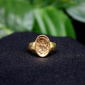 Pode incluir: Um anel de ouro com um design oval que apresenta uma imagem detalhada da cabe&ccedil;a de um guerreiro de perfil, usando um capacete.