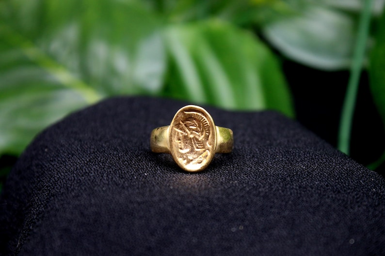 Pode incluir: Um anel de ouro com um design oval que apresenta uma imagem detalhada da cabe&ccedil;a de um guerreiro de perfil, usando um capacete.