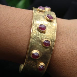 Pode incluir: Uma pulseira larga de punho dourada martelada, adornada com várias pedras preciosas ovais vermelho escuro. A pulseira tem uma superfície texturizada e é cravejada com as pedras preciosas em biséis de ouro individuais.
