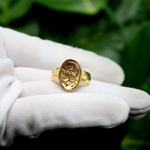 Pode incluir: Um anel de ouro com um design oval em relevo que apresenta o perfil de uma pessoa com capacete. O anel est&aacute; sobre uma superf&iacute;cie branca.