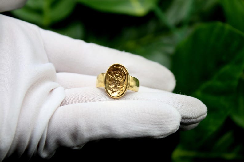 Pode incluir: Um anel de ouro com um design oval em relevo que apresenta o perfil de uma pessoa com capacete. O anel est&aacute; sobre uma superf&iacute;cie branca.