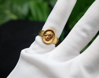May include: A gold ring with a round, raised design featuring a profile of a person's face.