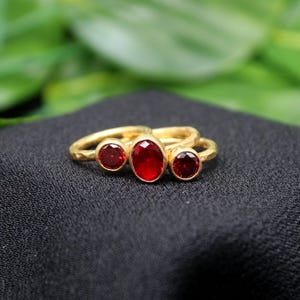 May include: Gold ring with three red gemstones. The center stone is oval shaped and the two side stones are round.