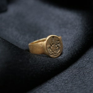 May include: A gold signet ring with an intricate design on the face. The ring is on a black fabric background.