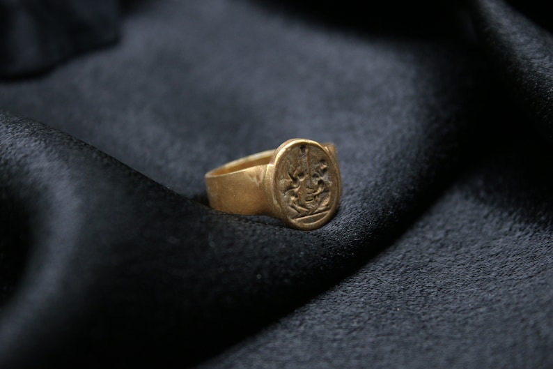 May include: A gold signet ring with an intricate design on the face. The ring is on a black fabric background.
