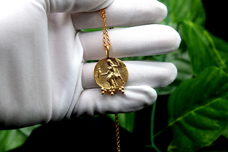 May include: A gold necklace with a round pendant featuring a detailed relief of a woman riding a horse. The pendant has five small gold beads hanging from the bottom.