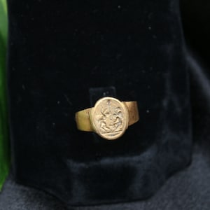 May include: A gold ring with an oval-shaped setting featuring a detailed engraved design. The ring is displayed on a black velvet background.