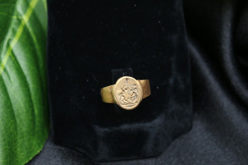 May include: A gold ring with an oval-shaped setting featuring a detailed engraved design. The ring is displayed on a black velvet background.