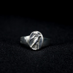 Pu&ograve; includere: Un anello d'argento con un designo in rilievo di una donna con un vestito fluido. L'anello &egrave; su uno sfondo nero.